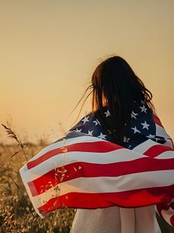 A young girl stands in a sunlit field, with an American flag draped over her shoulders
