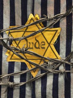 Yellow Star of David badge marked “Jude” pinned to a striped concentration camp uniform, with strands of barbed wire crossing the foreground.