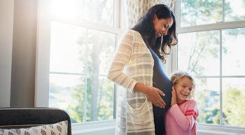 a pregnant woman smiling as a young girl leans against her belly, listening with excitement