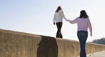 A woman helps a young girl balance while walking along the top of a concrete wall under a clear sky.