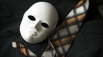 A blank white face mask rests on a black suit beside a gold, gray, and black plaid necktie.