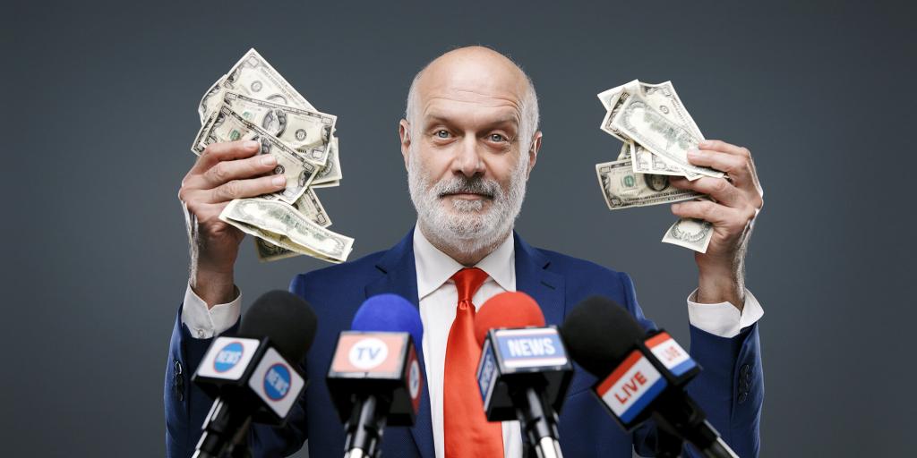 politician standing in front of reporters microphones and holding wads of cash