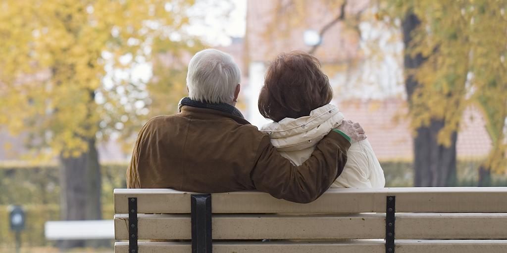 An elderly couple sitting on a park bench in Autumn with their backs to the viewer