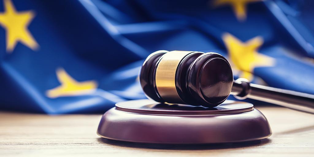 A gavel resting on a wooden block, with a defocused and rumpled E.U. flag draped on the table behind it.