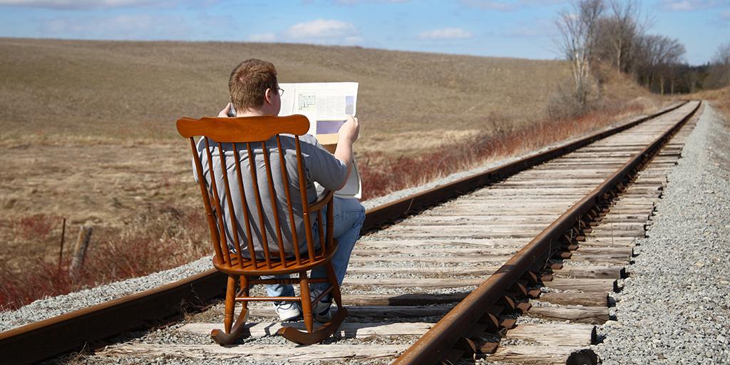 A man reading a periodical while sitting in a rocking chair in the middle of train tracks