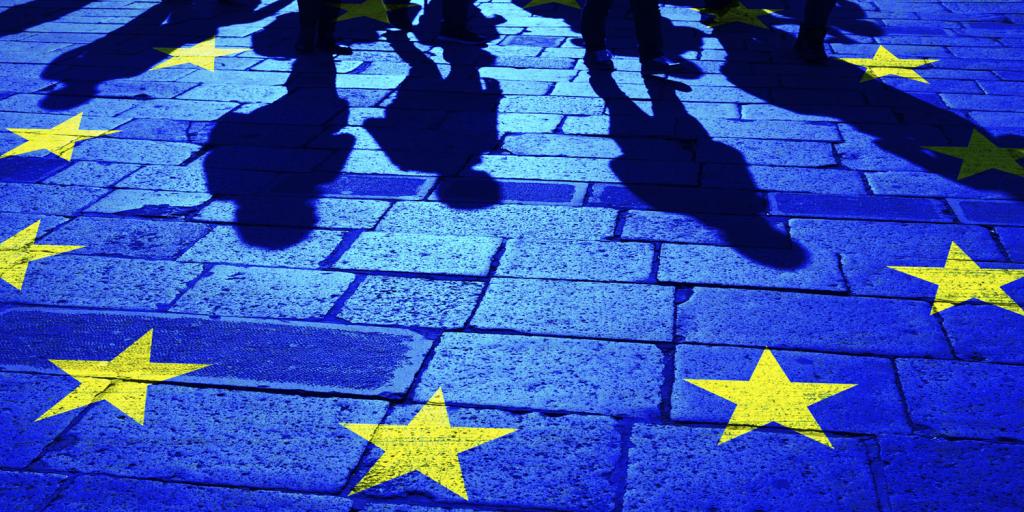cobblestone street with shadows of migrants approaching a circle of yellow stars representing the European Union