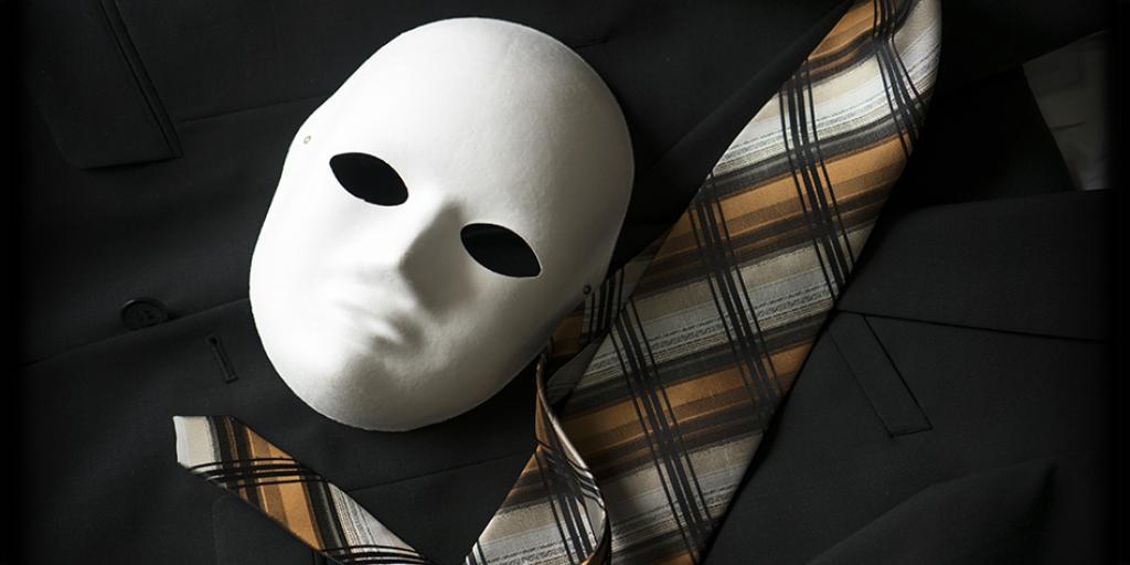 A blank white face mask rests on a black suit beside a gold, gray, and black plaid necktie.
