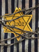 Yellow Star of David badge marked “Jude” pinned to a striped concentration camp uniform, with strands of barbed wire crossing the foreground.