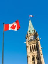 A large Canadian flag waves in the foreground, with the iconic Peace Tower of Parliament Hill in Ottawa visible behind it, topped by a smaller Canadian flag, all set against a clear blue sky