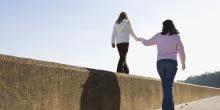 A woman helps a young girl balance while walking along the top of a concrete wall under a clear sky.